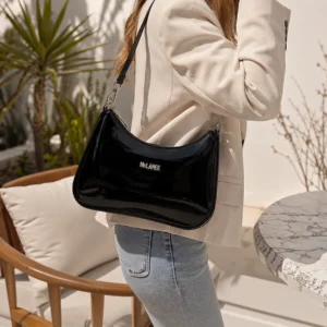 Black patent leather McLANEE Stirling hobo bag with a glossy curved silhouette, worn over the shoulder with a cream blazer and jeans.
